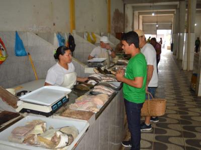 A imagem mostra um estudante conversando com uma vendedora no Mercado do Peixe