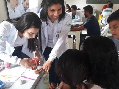 A imagem mostra duas alunos ensinando os pais e crianças a escovarem os dentes